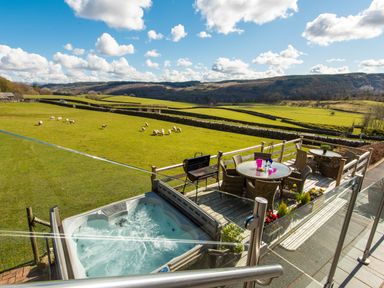 An outdoor patio with wicker chairs and tables a barbecue and a hot tub overlooking green fields with sheep and hills at Browside Little Arrow near Coniston