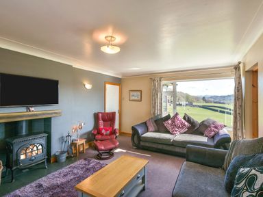 A living room with sofas a red armchair a wood stove and a large window showing fields at Browside Little Arrow near Coniston