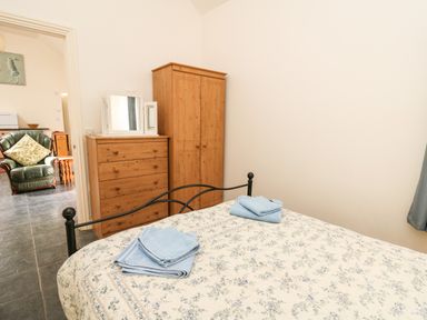A bedroom with a bed, folded blue towels, wooden chest of drawers with a mirror, wooden wardrobe, and an adjacent room with a green armchair at Yr Hen Ysgubor in Trelech near St Clears