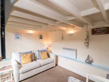 A living room with a beige sofa with patterned cushions a side table with books framed pictures on the wall and a wall heater at Nutshell in Whitby