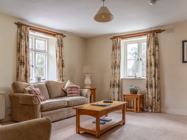 A sitting room with a beige sofa two windows with floral curtains wooden coffee and side tables with lamps and a floral basket at Crabtree Hawkshead