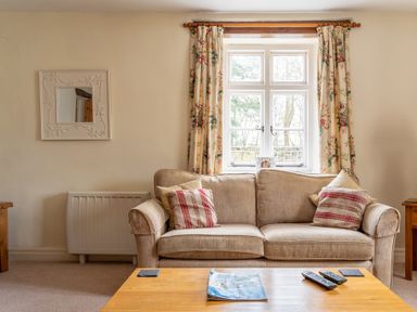 A beige sofa with patterned cushions in front of a window with floral curtains and a wooden coffee table with remotes and a magazine in Crabtree Hawkshead