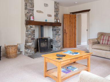 A living room with a wooden coffee table remote controls and books a stone fireplace and a beige sofa at Crabtree in Hawkshead