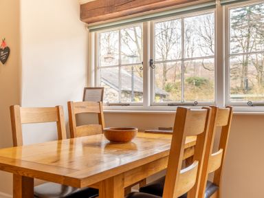 A wooden dining table with four wooden chairs and a bowl on the table by a window with a view of trees and a building at Crabtree in Hawkshead