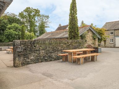 An outdoor seating area with wooden benches and tables beside a stone wall and buildings at Forge in Graythwaite near Hawkshead