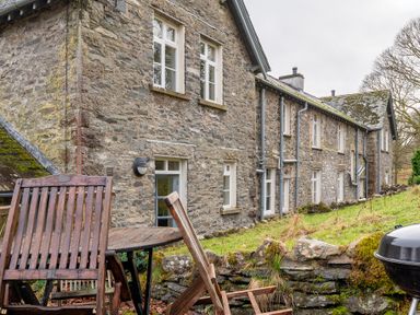 A stone house with multiple windows and a grassy yard with wooden chairs and a table at Forge in Graythwaite near Hawkshead