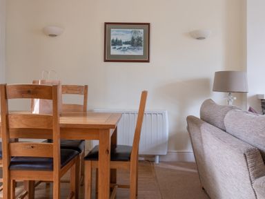 A dining area with a wooden table and chairs a paper bag on the table a framed painting on the wall wall mounted lights a beige sofa and a table lamp at Forge in Graythwaite near Hawkshead