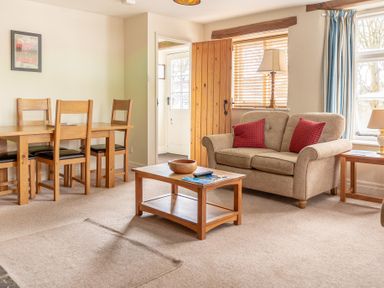 A living room with a wooden dining table and chairs a sofa with red cushions a coffee table and windows with blue curtains at Newlands Graythwaite near Hawkshead