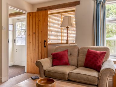 A living room with a beige sofa with red cushions a wooden coffee table a lamp by the window and a wooden door at Newlands in Graythwaite near Hawkshead