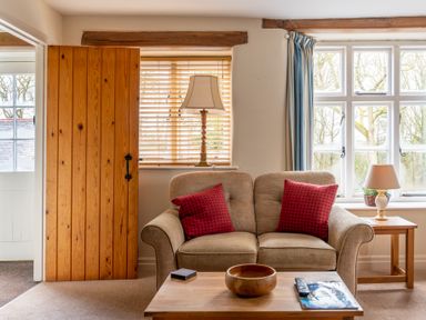 A living room with a beige sofa with red cushions a wooden coffee table with a bowl and a remote control a wooden side table with a lamp and plant wooden blinds and windows with blue curtains at Newlands in Graythwaite near Hawkshead