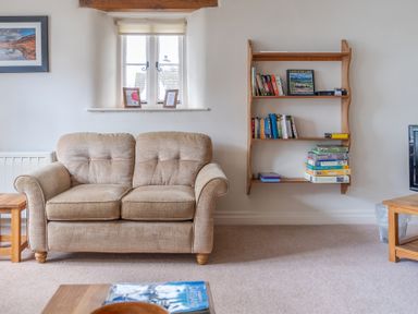 A beige sofa under a window with two picture frames a wooden bookshelf with books and board games and a wooden side table at Newlands Graythwaite near Hawkshead