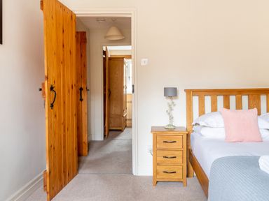 A bedroom with a wooden bed and bedside table with a lamp and an open door showing a hallway with wooden wardrobes at Newlands Graythwaite near Hawkshead