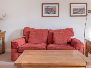 A red sofa with cushions against a white wall with two framed landscape pictures a wooden coffee table in front at Greenhowes Graythwaite near Hawkshead