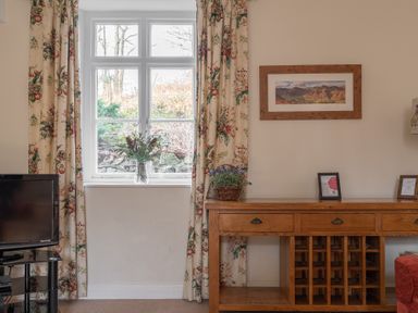 A room with a window with floral curtains a wooden sideboard with drawers and baskets and a television on a black stand at Greenhowes in Graythwaite near Hawkshead