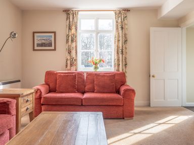 A living room with a red sofa a wooden coffee table a side table flowers on the windowsill and a door at Greenhowes in Graythwaite near Hawkshead