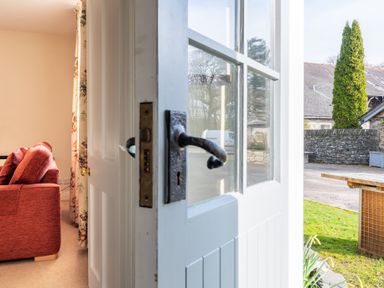 An open white door with glass panels showing a red sofa inside and a garden with a stone wall outside at Greenhowes in Graythwaite near Hawkshead