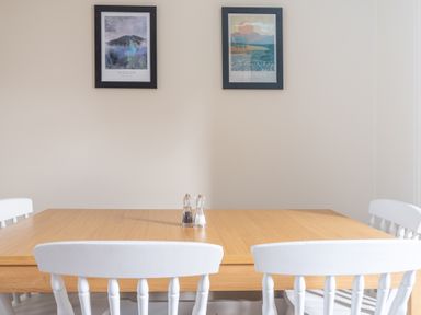 A dining area with a wooden table and four white chairs with two framed picture prints on the wall at Greenhowes Graythwaite near Hawkshead