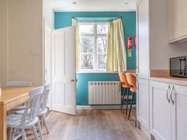 A kitchen with white cabinets a wooden dining table with white chairs a window with yellow curtains and two orange bar stools at Greenhowes Graythwaite near Hawkshead