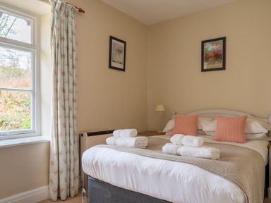 A bedroom with a bed with folded towels two pink cushions a bedside table with a lamp and floral curtains at Greenhowes Graythwaite near Hawkshead
