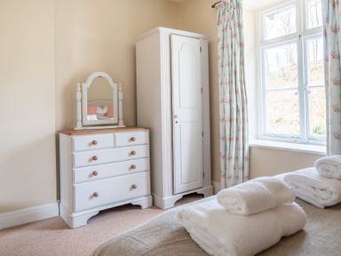 A bedroom corner with a white dresser with a mirror and a wardrobe next to a window with floral curtains at Greenhowes Graythwaite near Hawkshead