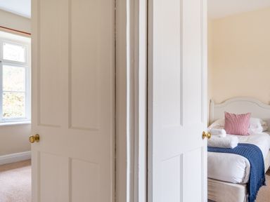 A bedroom with a single bed with white linens a blue throw and a pink pillow seen through an open door at Greenhowes Graythwaite near Hawkshead