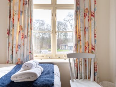 A bedroom window with floral curtains a white chair and towels on a bed at Greenhowes Graythwaite near Hawkshead