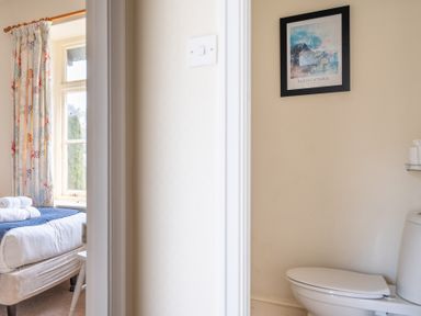 A bathroom with a toilet and a framed picture on the wall next to a bedroom with a bed, a window, and floral curtains at Greenhowes Graythwaite near Hawkshead