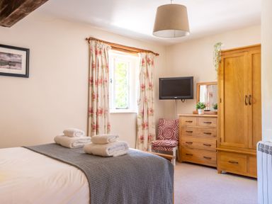 A bedroom with a bed, folded towels, floral curtains, wooden wardrobe, dresser with a mirror, chair, and a wall-mounted television at Lord's Leap in Graythwaite near Hawkshead