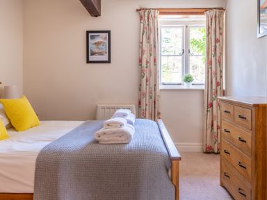 A bedroom with a bed with yellow pillows and gray blanket near a window with floral curtains and a wooden dresser at Lords Leap Graythwaite near Hawkshead