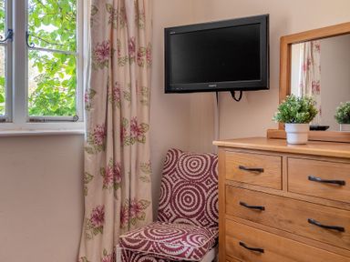 A wooden dresser with drawers and a mirror with a small potted plant on top next to a patterned chair and window with floral curtains at Lords Leap Graythwaite near Hawkshead