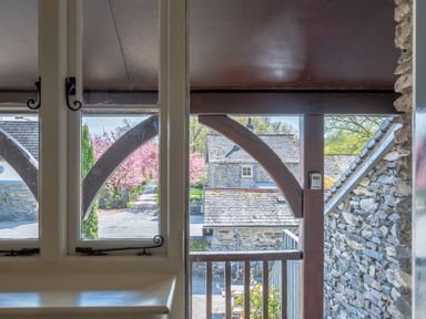 View through a window and wooden porch structure to stone buildings and a path lined with pink blossoming trees at Lords Leap in Graythwaite near Hawkshead