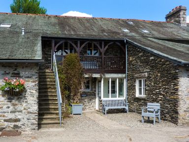 A stone exterior with stairs on the left flowers in a wall planter two blue benches and a blue armchair at Bassicks in Hawkshead