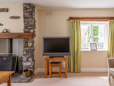 A living room with a stone fireplace wooden tables a television and green patterned curtains at Bassicks in Hawkshead