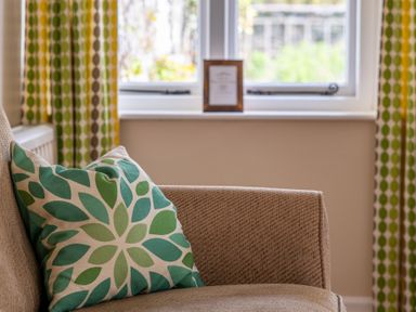A beige armchair with a green patterned cushion near a window with green and yellow curtains at Bassicks in Hawkshead