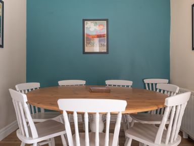 A dining room with a round wooden table and six white chairs with a framed picture on a teal wall at Bassicks in Hawkshead