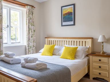 A bedroom with a bed with yellow cushions and rolled towels on it next to a nightstand with a lamp and a window with floral curtains at Bassicks in Hawkshead