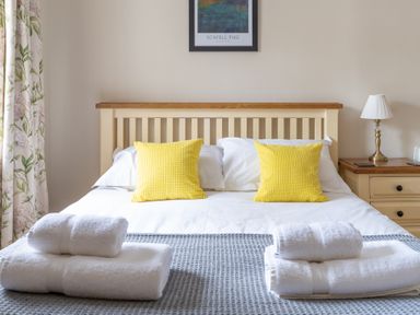 A bedroom with a bed with yellow pillows white towels on a grey blanket a bedside table with a lamp and floral curtains at Bassicks Hawkshead