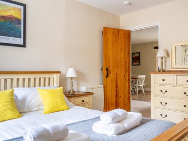 A bedroom with a bed with yellow pillows and folded towels on it a wooden dresser with a lamp and an open wooden door showing a dining area at Bassicks in Hawkshead