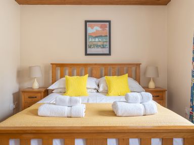 A bedroom with a wooden bed frame two yellow pillows folded white towels on a yellow bedspread two wooden bedside tables with lamps and a colorful picture on the wall at Bassicks in Hawkshead