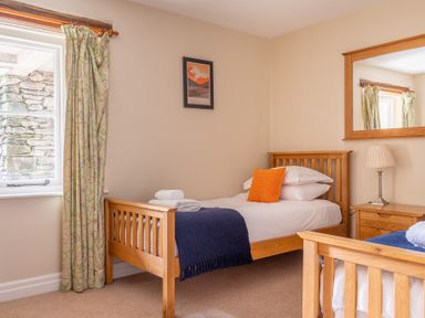 A bedroom with two single wooden beds with white bedding and blue throws a bedside table with a lamp a window with floral curtains and a framed picture on the wall at Bassicks in Hawkshead