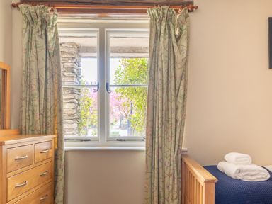 A bedroom window with floral curtains beside a wooden chest of drawers and a bed with folded towels at Bassicks in Hawkshead