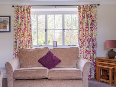 A beige sofa with a purple cushion in front of a window with floral curtains and a wooden side table with a lamp