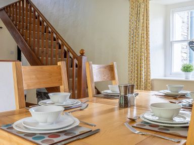 A dining table set with white bowls and plates near a wooden staircase and window with patterned curtains at Hilltop in Graythwaite near Hawkshead