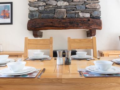 A dining table set with white bowls plates silverware and salt and pepper shakers in front of a stone and wood fireplace at Hilltop in Graythwaite near Hawkshead