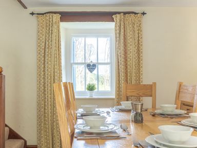 A dining room with a wooden table set with plates bowls cutlery and glasses next to a window with patterned curtains at Hilltop in Graythwaite near Hawkshead