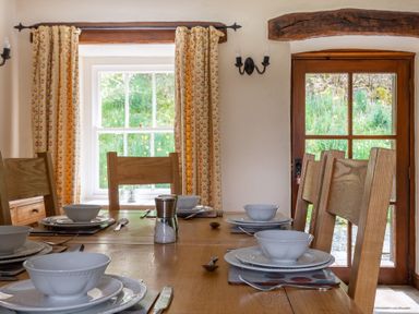 A dining room with a wooden table set with white bowls plates and cutlery near windows with patterned curtains at Hilltop in Graythwaite near Hawkshead