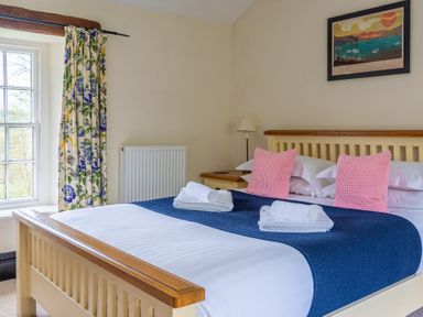 A bedroom with a bed with pink pillows and a blue blanket near a window with floral curtains at Hilltop in Graythwaite near Hawkshead