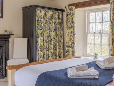 A bedroom with a bed covered with white and blue linens overlooking a window with floral curtains at Hilltop in Graythwaite near Hawkshead
