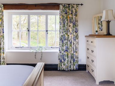 A bedroom window with floral curtains beside a white dresser with a mirror and lamp at Hilltop in Graythwaite near Hawkshead