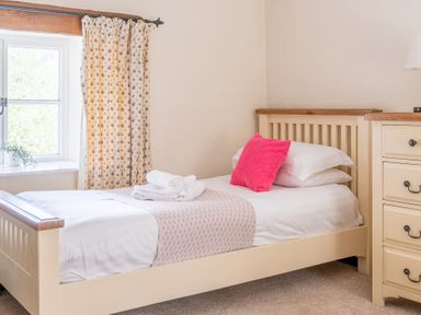 A single bed with white linens and a pink pillow next to a window with floral curtains and a cream dresser in a bedroom at Hilltop in Graythwaite near Hawkshead